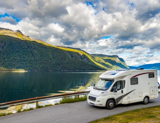 An RV camper driving along a curved road on a mountain. The road overlooks a lake and there's a blue cloudy sky above.