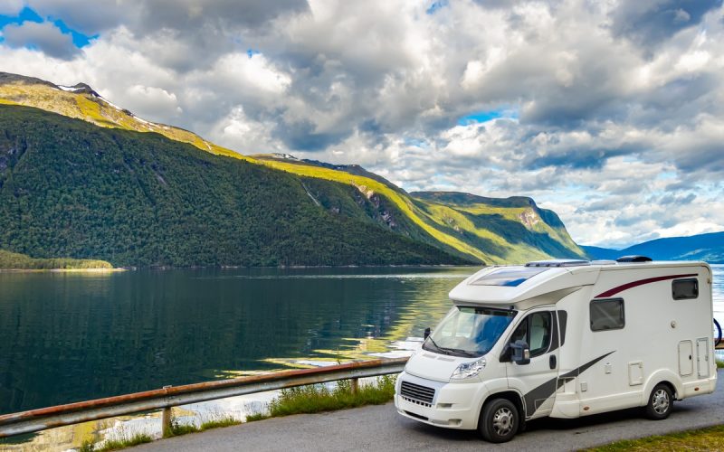 An RV camper driving along a curved road on a mountain. The road overlooks a lake and there's a blue cloudy sky above.