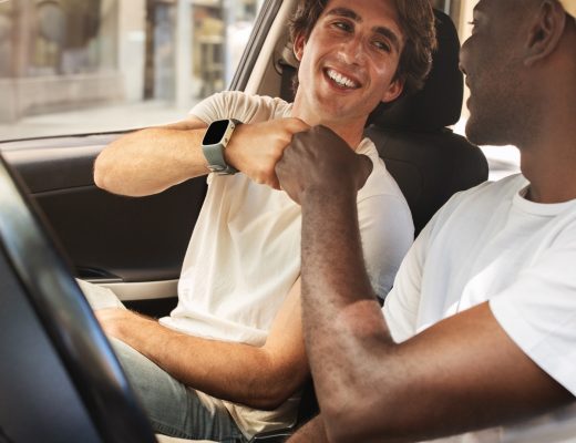 Two smiling men in white t-shirts sit in the front seats of a parked car and give each other a fist bump.