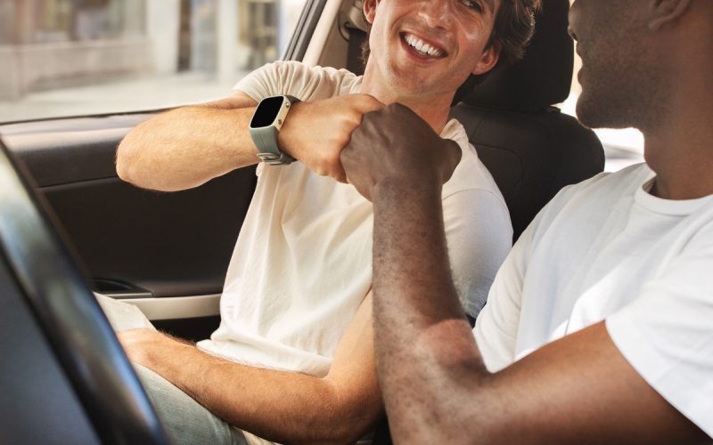 Two smiling men in white t-shirts sit in the front seats of a parked car and give each other a fist bump.