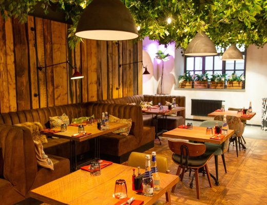 The interior of a dimly lit contemporary restaurant with upholstered booths, small square tables, and vines on the ceiling.