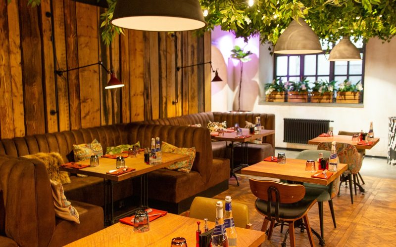The interior of a dimly lit contemporary restaurant with upholstered booths, small square tables, and vines on the ceiling.