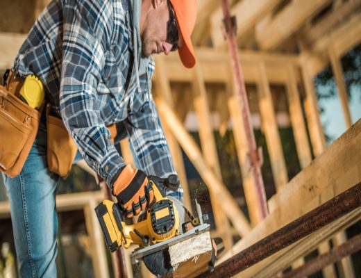 A man wearing an orange hat, sunglasses, and a plaid shirt is using a large yellow power tool on a piece of wood.