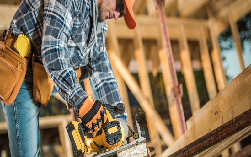 A man wearing an orange hat, sunglasses, and a plaid shirt is using a large yellow power tool on a piece of wood.