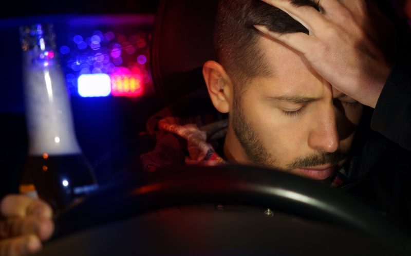 A man sits in the driver's seat of a car while holding a bottle in one hand and resting his head in the other hand.
