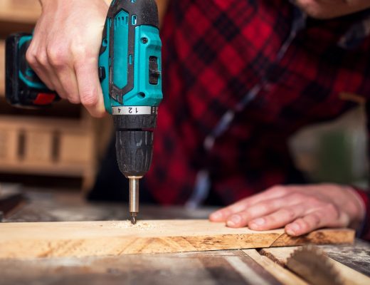 A close-up of a man in a black and red flannel shirt using a power tool to drill a piece of wood in a workshop.