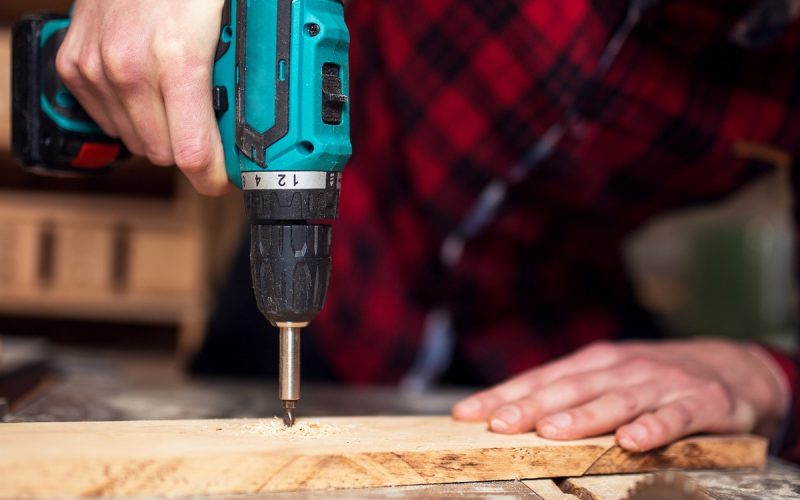 A close-up of a man in a black and red flannel shirt using a power tool to drill a piece of wood in a workshop.