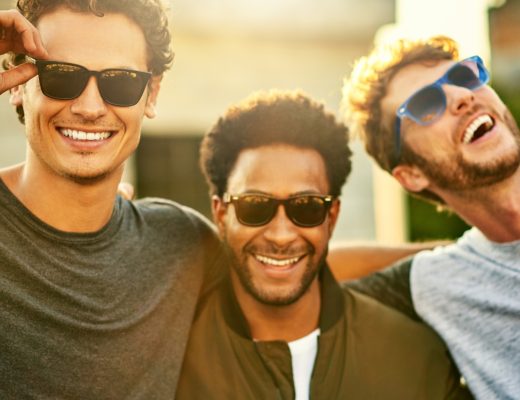 A group of three guy friends standing shoulder to shoulder outside in sunglasses. They're all smiling.