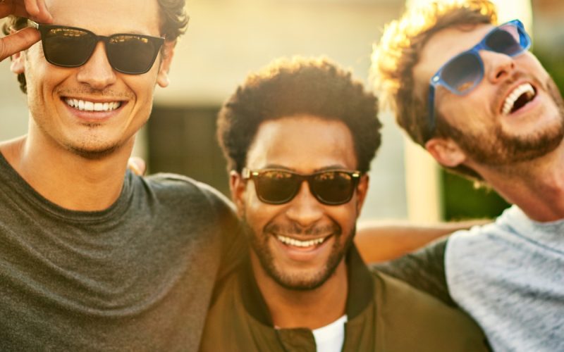 A group of three guy friends standing shoulder to shoulder outside in sunglasses. They're all smiling.