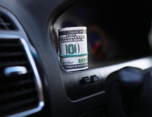 A roll of money with a visible $100 bill on the outside sitting on the clean black dashboard of a vehicle.
