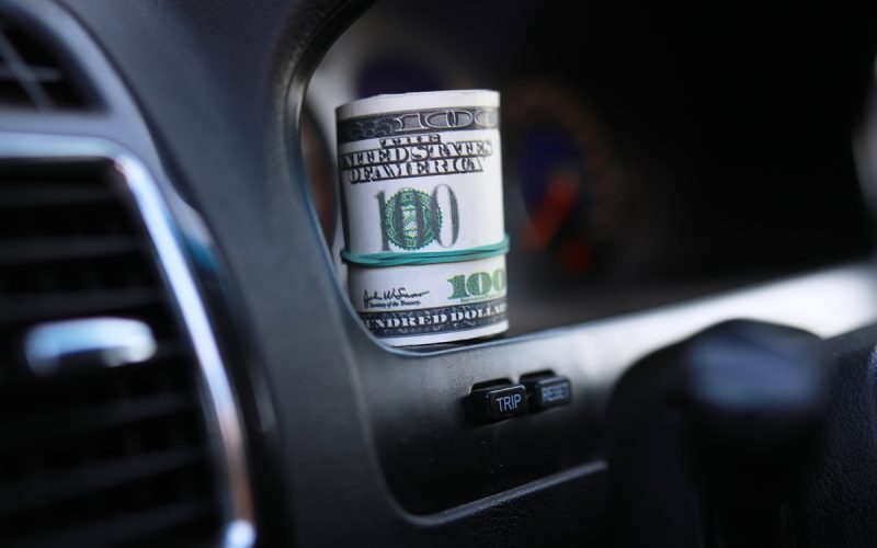 A roll of money with a visible $100 bill on the outside sitting on the clean black dashboard of a vehicle.