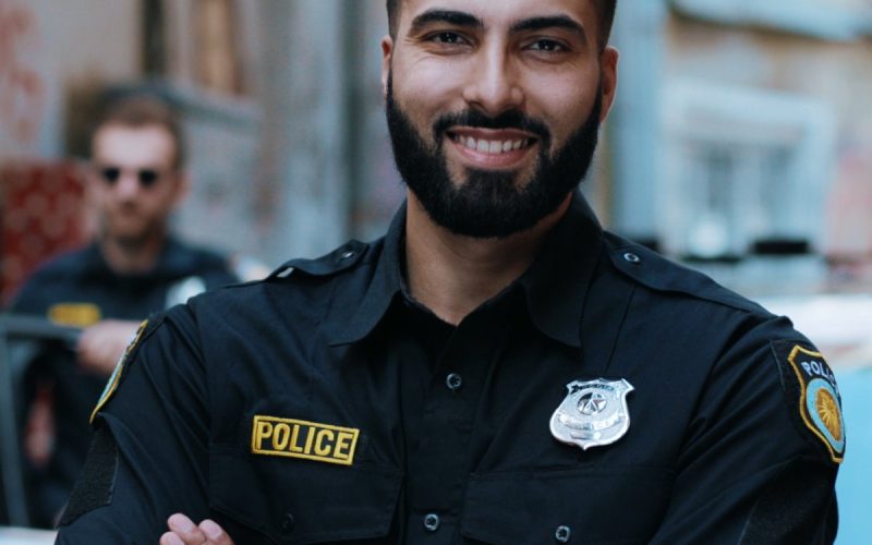 A police officer standing in front of his squad car with his arms crossed. His partner is standing outside the door behind him.