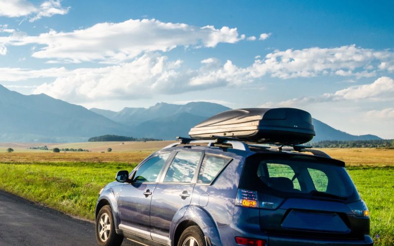 A hatchback car with storage on top of the hood is parked in the middle of the road in front of a mountainous view.