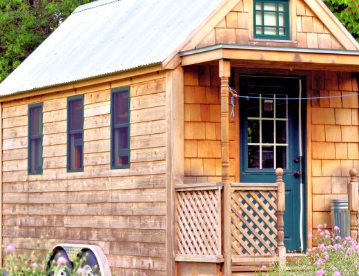 The exterior of a tiny wooden house on wheels. The house has a porch, several small windows, and a metal roof.