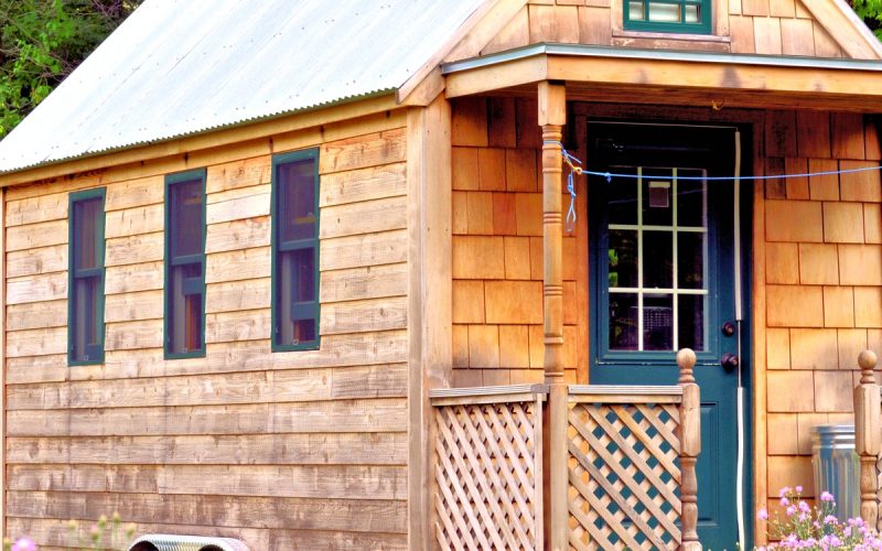The exterior of a tiny wooden house on wheels. The house has a porch, several small windows, and a metal roof.