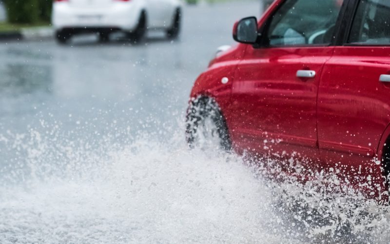 A red car is driving through a flooded zone in a residential area. There is a white vehicle in front of the red car.
