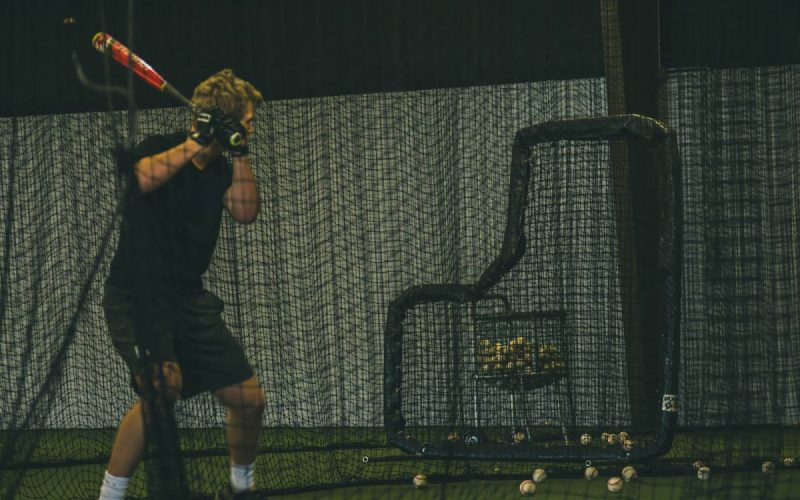 A young man in a batting stance is practicing hitting inside a batting cage surrounded by nets and baseballs.
