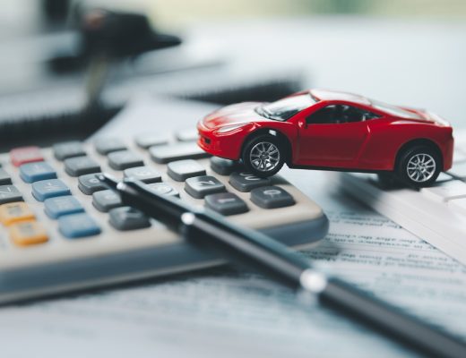 A red plastic model car resting on top of two calculators. A blurred pen, document, and laptop also occupy the scene.