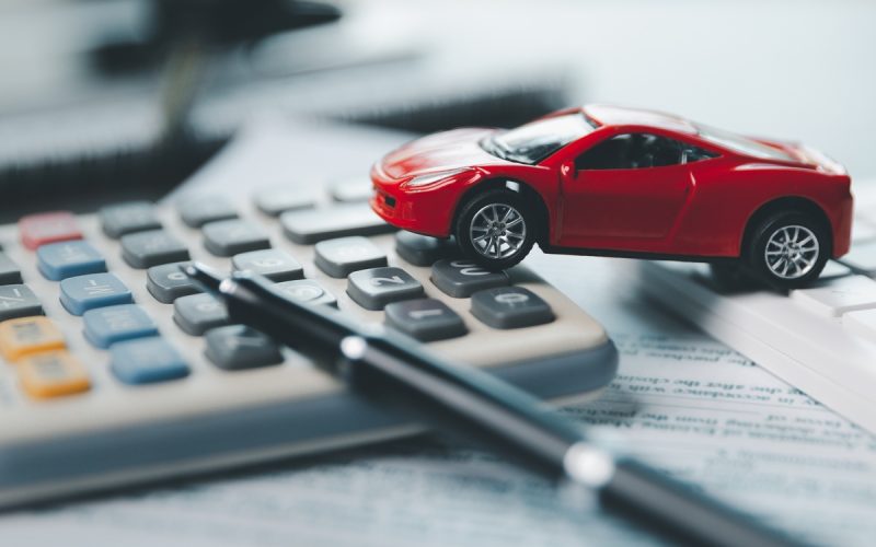 A red plastic model car resting on top of two calculators. A blurred pen, document, and laptop also occupy the scene.