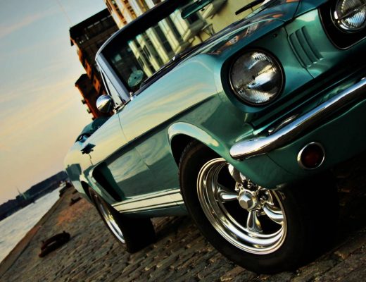 A vintage teal convertible with a boxy frame parked on a cobblestone street next to a river at sunset.
