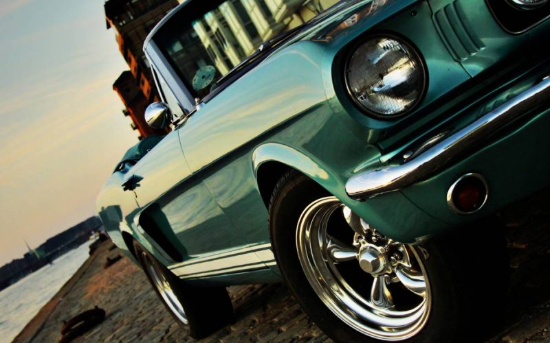 A vintage teal convertible with a boxy frame parked on a cobblestone street next to a river at sunset.