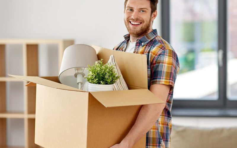 A man smiles and holds a box with a lamp and houseplant in a living room. A loveseat and empty bookshelf are behind him.