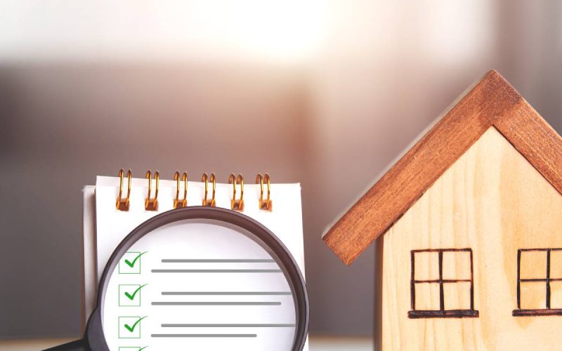 A magnifying glass hovering over a notepad with green check marks and gray lines, next to a wooden house.