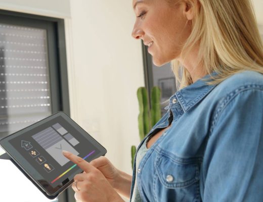 A woman standing in front of a sliding glass door. She is using an electronic tablet to operate rolling shutters over the door.