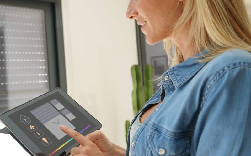 A woman standing in front of a sliding glass door. She is using an electronic tablet to operate rolling shutters over the door.