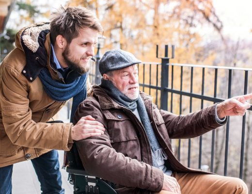 A son walking outside with his older father, who is in a wheelchair. They are both wearing winter attire.