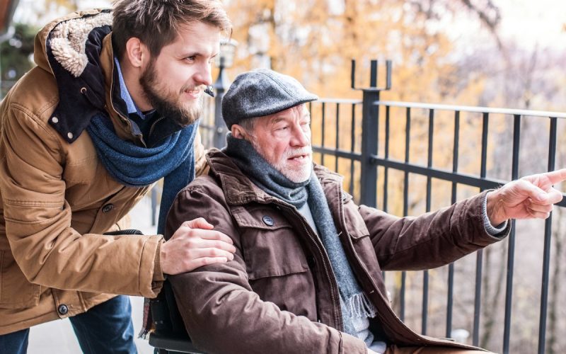 A son walking outside with his older father, who is in a wheelchair. They are both wearing winter attire.
