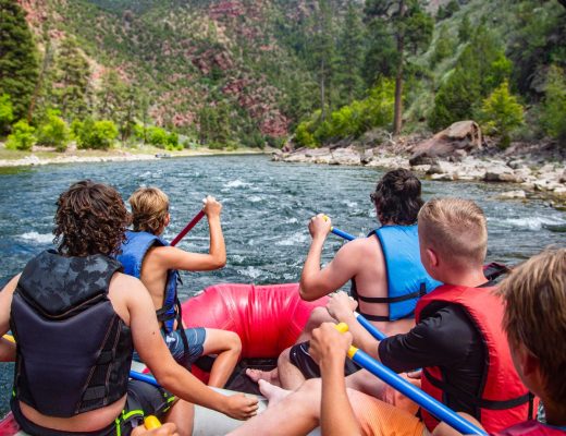 A large group of men is on a white water rafting trip down a rapid river. They're all wearing red life vests.