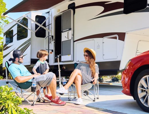 Young parents sits on chairs outside near camping trailer and car while talking to their school-aged son.