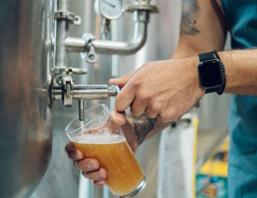 A close-up of a person filling a glass of beer from a tap in a brewery. They're wearing a blue apron and an Apple Watch.