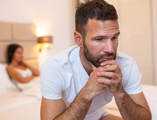A man sits on the edge of a bed with a frustrated expression. A woman sits on the other side of the bed under the covers.