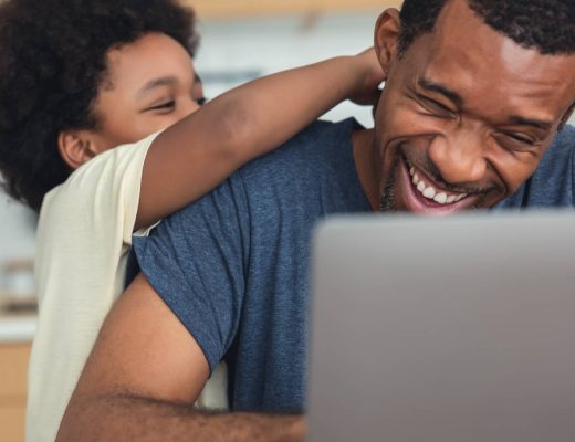A father working from home on his laptop in his kitchen laughs as his son tickles the back of his head.