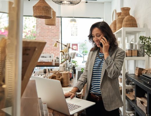 A woman wearing a blazer stands behind the counter at her business. She talks on the phone while she works on her laptop.