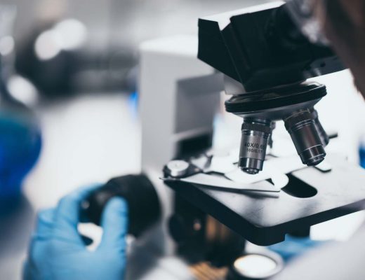 A young male scientist wearing blue gloves changes the focus on a microscope that he looks into to see a slide.