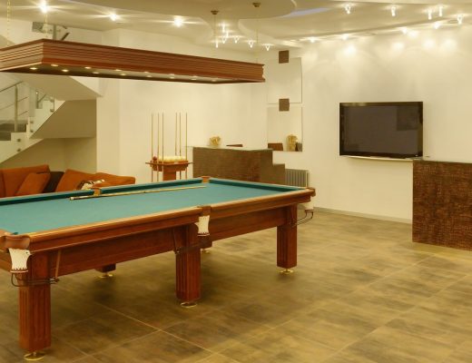 A billiards table sits on a tiled floor between an orange couch and a table with brown chairs in a basement.