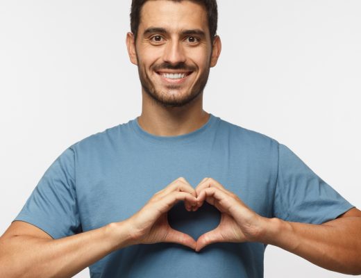 A smiling man makes a heart symbol with his hands and holds it over his chest. He stands in front of a gray wall.