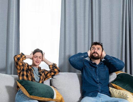 A man and a woman sit on their living room sofa, holding their hands over their ears to block out noise.