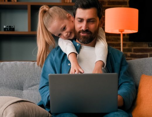 A man sits on a gray couch with a laptop placed on his lap. A blonde child behind him reaches toward the laptop.