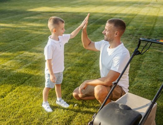 A boy in a white shirt standing on the grass high-fives a man in a white shirt kneeling on the grass next to a lawnmower.