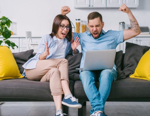 A man and woman sit on their couch in their home. They cheer excitedly as they win an item on an online auction.