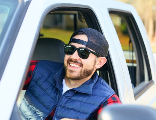 A man wearing a backwards cap and sunglasses smiles and leans out the side window of a white pickup truck.