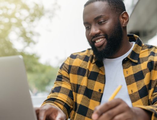 A young Black man is using a laptop, taking notes and planning, while smiling and sitting outside.