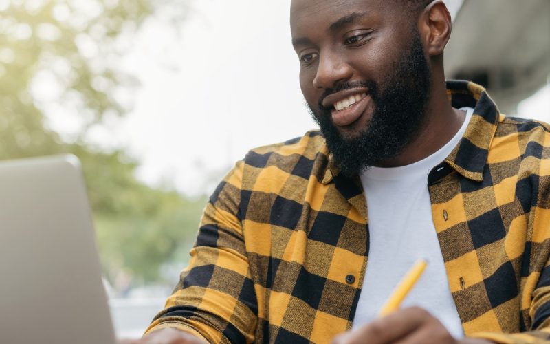 A young Black man is using a laptop, taking notes and planning, while smiling and sitting outside.