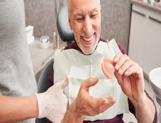 A smiling senior man in a maroon turtleneck holds a partial set of dentures while sitting in a dentist's chair.