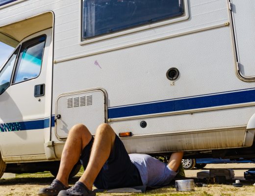 A man lies beneath an RV, repairing it with tools nearby. The RV is parked outdoors on a patchy grass surface.