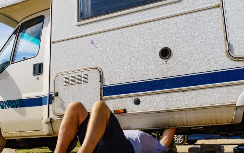 A man lies beneath an RV, repairing it with tools nearby. The RV is parked outdoors on a patchy grass surface.